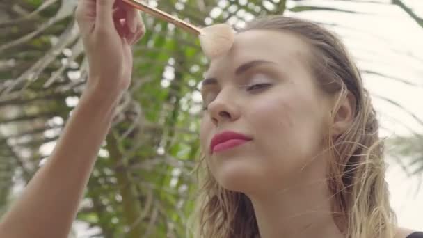 Gros plan du mannequin recevant du maquillage pendant la séance photo de la plage, maquilleuse ajustant le fard à joues sur son visage avec un pinceau entre les palmiers — Video