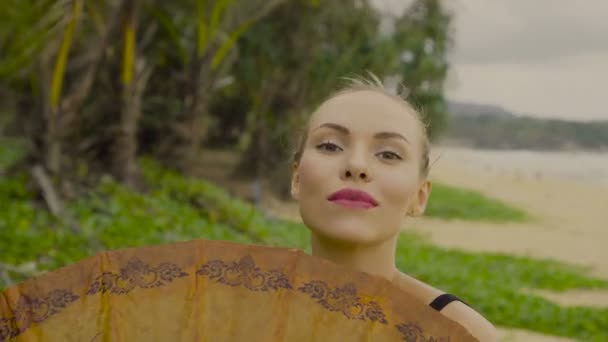 Belle femme blonde glamour portant des maillots de bain noirs avec parapluie chinois souriant et posant par une belle journée d'été sur fond de plage et de palmiers — Video