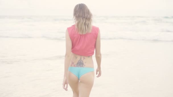 Widok z tyłu Piękna Blondynka seksowna kobieta ubrana w czerwoną koszulkę i niebieskie bikini dno chodzenie do morza na pięknym letnim dniu-wideo w zwolnionym tempie — Wideo stockowe