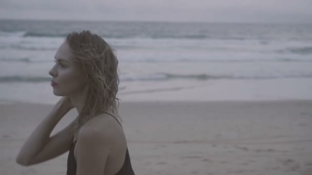 Schöne blonde Glamour-Frau in schwarzem Kleid, die an einem Spätsommernachmittag an einem Strand spaziert, mit nassen Haaren über Meer und Himmel -Video in Zeitlupe — Stockvideo