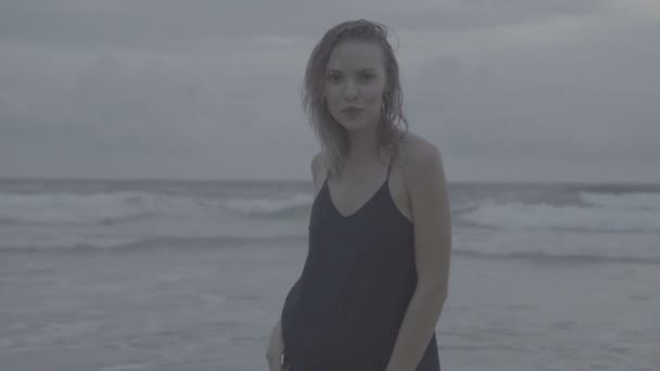 Linda mulher loira glamour vestindo vestido preto posando em uma praia no final da tarde de verão com cabelo molhado sobre o fundo do mar e do céu — Vídeo de Stock