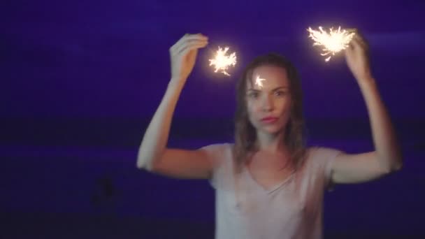 Primer plano de la sensual mujer que lleva camiseta con el pelo mojado haciendo fuegos artificiales chispeantes muestran en la playa por la noche sobre el mar y el fondo del cielo — Vídeo de stock