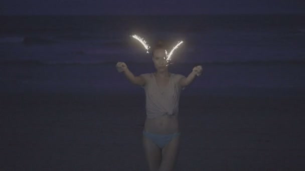 Sensual mujer con camiseta y parte inferior del bikini haciendo fuegos artificiales más brillantes muestran en la playa por la noche sobre el mar y el fondo del cielo — Vídeos de Stock