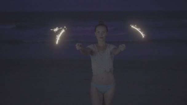 티셔츠와 비키니 하의를 입은 관능적인 여성이 바다와 하늘을 배경으로 밤에 해변에서 반짝이는 불꽃 놀이쇼를 합니다. — 비디오