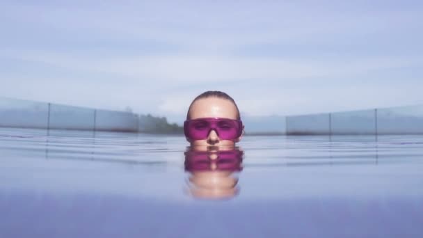 Primer plano de la cara de la mujer joven sexy con gafas de sol púrpura mirando a la cámara en la piscina infinita en la azotea en un día soleado sobre el cielo azul — Vídeo de stock