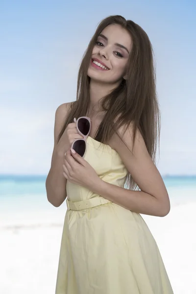 Νεαρή και ευτυχισμένη γυναίκα με κίτρινο φόρεμα που ποζάρει με γυαλιά ηλίου στο χέρι στην παραλία. Νιώθω ελεύθερος και χαρούμενος — Φωτογραφία Αρχείου