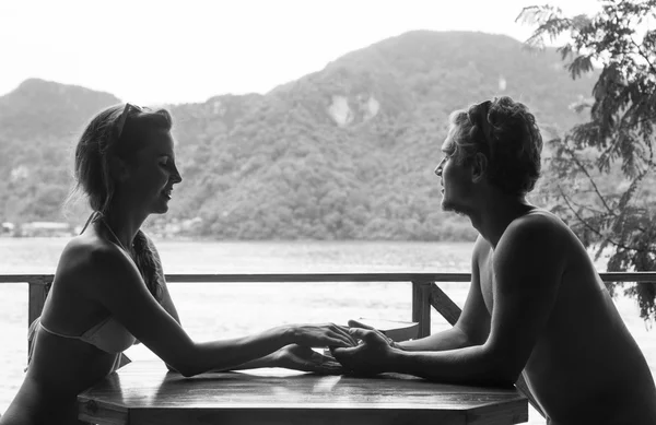 Χαριτωμένο ζευγάρι φορώντας μαγιό στην αγάπη κρατώντας τα χέρια και χαμογελώντας κάθεται στο τραπέζι σε φιλόξενο καλοκαιρινό καφέ στην παραλία — Φωτογραφία Αρχείου