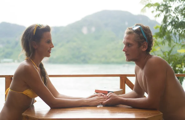 Couple mignon portant des maillots de bain en amour tenant la main et souriant assis à la table dans un café d'été confortable sur la plage — Photo