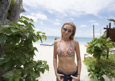 Gülümseyen güzel sarışın bikini sütyen giyen, şort ve sungalsses deniz ve gökyüzü arka plan üzerinde plajda