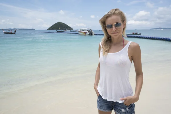 Leende Söt blondin bär jeansshorts, vit t-shirt och solglasögon på stranden över hav och himmel bakgrund — Stockfoto