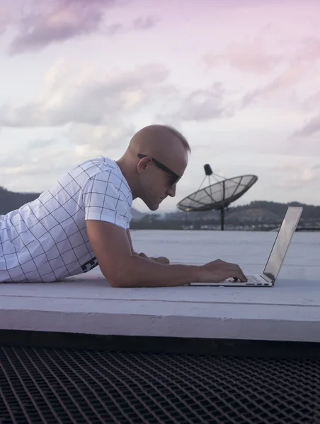 Gün batımı gökyüzü arka plan üzerinde bir çatı üzerinde yatan dizüstü bilgisayar ile güneş gözlüğü yakışıklı genç adamın Outdoors portre. Serbest ve teknoloji — Stok fotoğraf