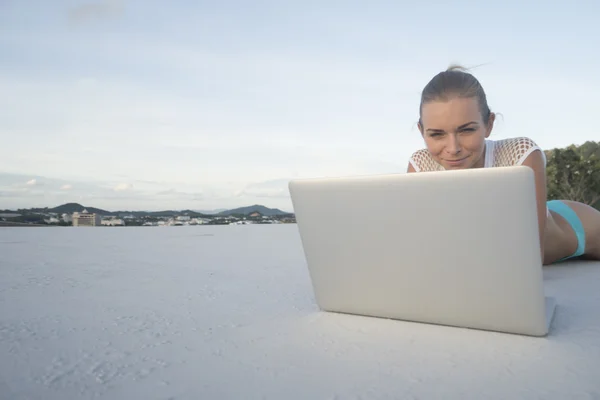 Außenporträt einer hübschen blonden Frau im Bikini mit Laptop, die auf einem Dach über der Stadtlandschaft und dem Himmel liegt. freiberuflich und technologisch tätig — Stockfoto