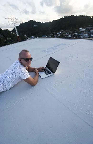 Outdoors portrait of handsome young man in sunglasses with laptop computer lying on a rooftop over citylandscape background. Freelance and technology — Stock Photo, Image