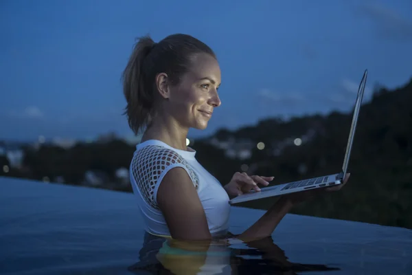 All'aperto ritratto di bella donna bionda felice con computer portatile in una piscina a sfioro sul tetto su sfondo sfocato montagna e cielo durante la sera. Freelance e tecnologia — Foto Stock