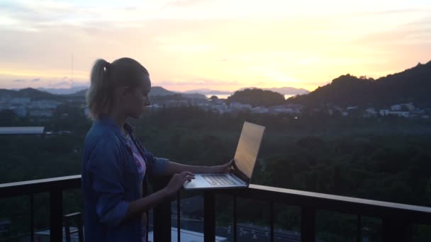 Vista lateral de la bonita mujer rubia en bikini y camisa jeans trabajando en una computadora portátil mientras está de pie en una azotea sobre el hermoso amanecer. Independiente y tecnología — Vídeos de Stock