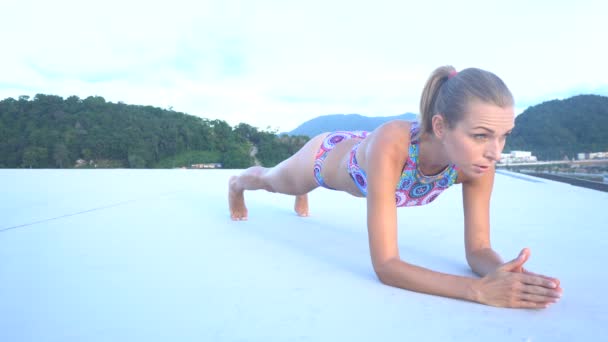 Botten sida vy över ponny tailed ganska blond kvinna klädd i färgglada Bikini gör plank push up övning på en takterrass över blå himmel och gröna bergstoppar under soluppgången. Sport och hälsosamt livsstilskoncept — Stockvideo