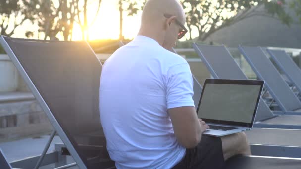 Portrait en plein air d'un beau jeune homme en lunettes de soleil travaillant sur un ordinateur portable assis sur un transat sur un toit au-dessus d'un beau lever de soleil. Freelance et technologie — Video