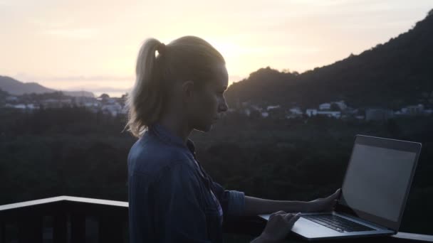 Vue latérale d'une jolie femme blonde en soutien-gorge de bikini et chemise en jean travaillant sur un ordinateur portable tout en se tenant sur un toit au-dessus d'un beau lever de soleil. Freelance et technologie — Video