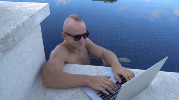 Открытый портрет красивого счастливого мужчины в солнечных очках, работающего на ноутбуке в бассейне на крыше. Фриланс и технологии — стоковое видео