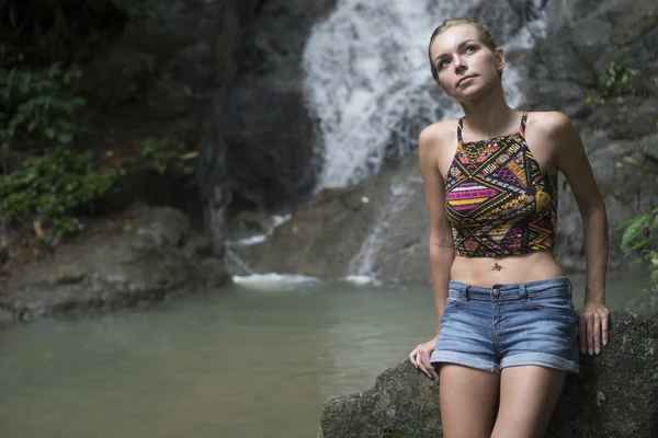 Söt blond kvinna sittting på en sten och letar upp nära vattenfallet. Kvinnliga turister njuter av en vatten falla i skogen — Stockfoto