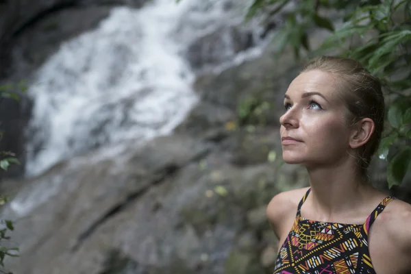 Primer plano de la cara de la hermosa mujer rubia mirando por encima de la cascada borrosa. Turista femenina disfrutando de una caída de agua en el bosque — Foto de Stock