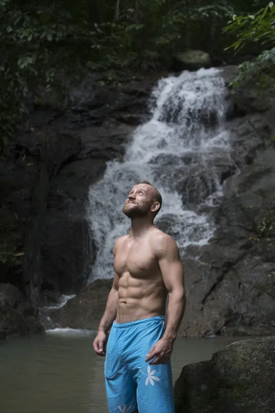 Hombre guapo con barba vistiendo pantalones cortos azules de pie y mirando hacia arriba cerca de la cascada. Turista masculino disfrutando de una caída de agua en el bosque — Foto de Stock