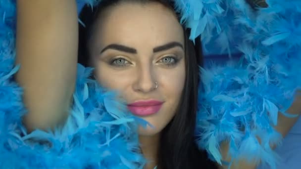 Primer plano de la hermosa mujer morena sonriendo y mirando a la cámara con boa de plumas azules sobre fondo de pared azul — Vídeo de stock