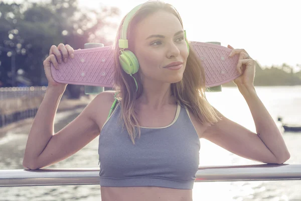 Ung vacker blond fitness kvinna bär ljusa gröna hörlurar lyssna musik och hålla rosa skateboard stående på bryggan nära havet, njuter av tidig morgon med vacker soluppgång — Stockfoto