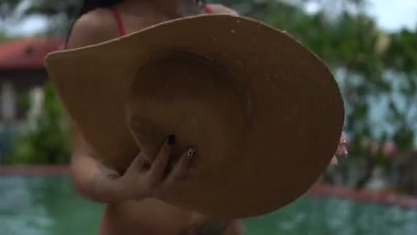 Beautiful sexy brunette woman wearing pink bikini putting on gold hat and relaxing by outdoor swimming pool at tropical resort — Stock Video