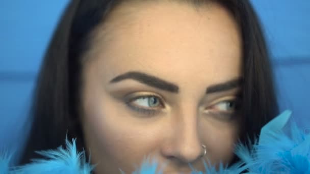 Close-up van de mooie brunette vrouw glimlachen en kijken in de camera met blauwe veren boa over blauwe muur achtergrond — Stockvideo