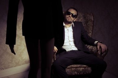 Genç ve zarif bir adam, siyah takım elbiseli, beyaz gömlekli ve güneş gözlüklü bir adam. Önünde duran kadına bakıyor. Geçmişe bakan kadın silueti, eski moda bir iç mekan giymiş yakışıklı bir erkeğin karşısında duruyor.