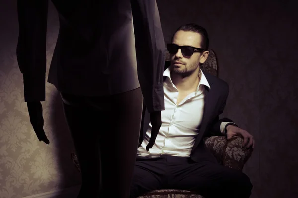 Young and elegant man sitting in chair wearing black suit, white shirt and sunglasses looking at woman standing in front of him isolated over vintage background. Back view silhouette of woman standing opposite handsome male in a retro inrerior — Stock Photo, Image