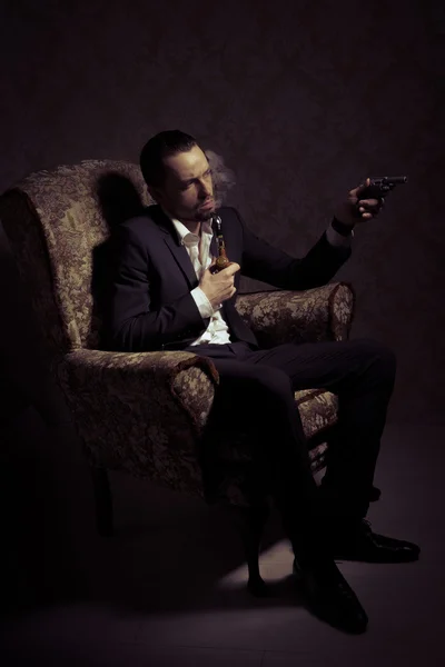 Young handsome and elegant man sitting in chair, holding gun and smoking a pipe isolated over vintage background — Stock Photo, Image