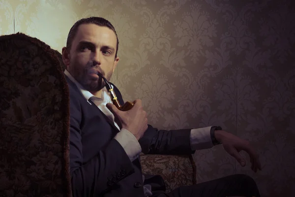 Jeune homme beau et élégant assis sur une chaise et fumant une pipe isolée sur fond vintage — Photo