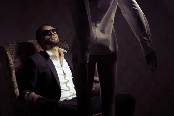 Hombre joven y elegante sentado en silla con traje negro, camisa blanca y gafas de sol mirando a la mujer de pie frente a él aislado sobre fondo vintage. Vista posterior silueta de la mujer de pie frente apuesto macho en un inrerior retro —  Fotos de Stock