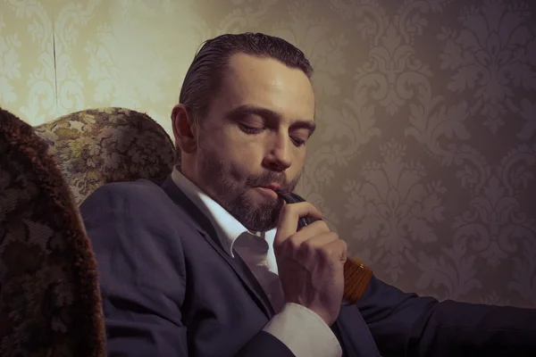 Jovem bonito e elegante homem sentado na cadeira e fumar um cachimbo isolado sobre fundo vintage — Fotografia de Stock
