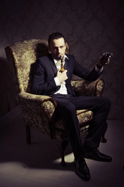 Jeune homme beau et élégant assis sur une chaise, tenant un pistolet et fumant une pipe isolée sur fond vintage — Photo