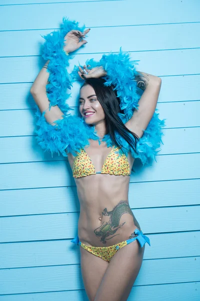 Hermosa mujer morena seductora con bikini amarillo y boa de plumas azules sonriendo y mirando a la cámara sobre fondo de pared de madera azul —  Fotos de Stock