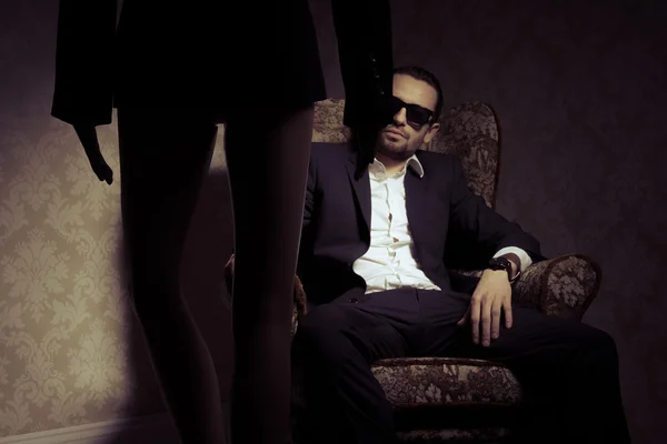 Young and elegant man sitting in chair wearing black suit, white shirt and sunglasses looking at woman standing in front of him isolated over vintage background — Stock Photo, Image