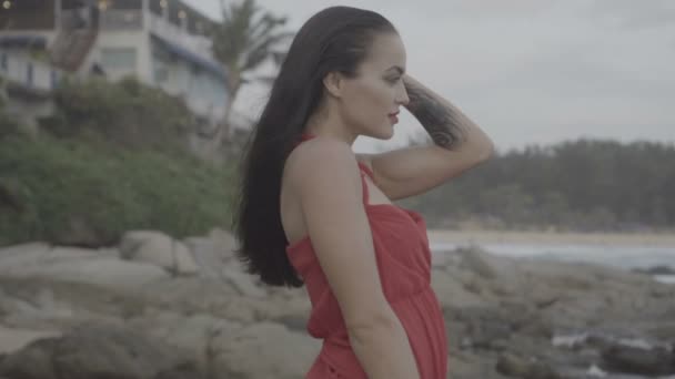 Piękna uwodzicielska kobieta w czerwonej sukience bawiąca się na skałach nad morzem — Wideo stockowe