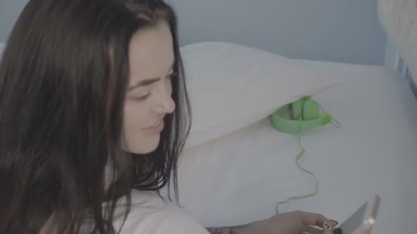 Όμορφη νεαρή μελαχρινή γυναίκα ξαπλωμένη στο κρεβάτι χρησιμοποιώντας το smartphone και τα ακουστικά της — Αρχείο Βίντεο