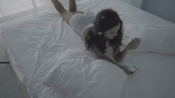 Bella giovane donna bruna sdraiata sul letto con il suo smartphone e le cuffie — Video Stock
