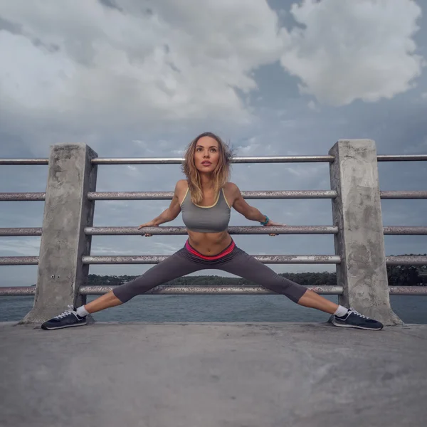 Junge hübsche blonde Frau in Trainingskleidung, die sich auf der Seebrücke in der Nähe des Meeres dehnt und den frühen Morgen mit schönem Sonnenaufgang genießt — Stockfoto