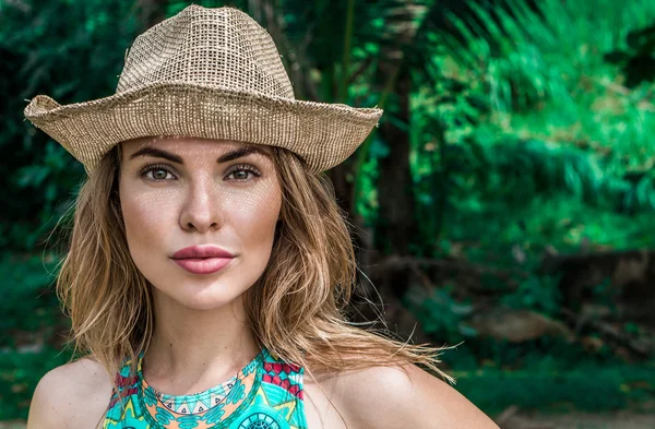 Retrato de bela jovem com chapéu de palha em um dia ensolarado olhando para a câmera sobre palmeiras fundo. Bonito olhos closeup de menina bonita vestindo top biquíni colorido e chapéu de palha sobre verde — Fotografia de Stock