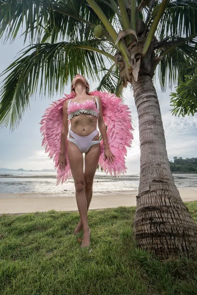 Romantic young beauty as an angel on the tropical beach looking up on the sky. Bottom view of blonde pretty female woman wearing pink wings, feather bra and bikini bottom standing on green grass close to palm tree over sea and sky background — Stock Photo, Image