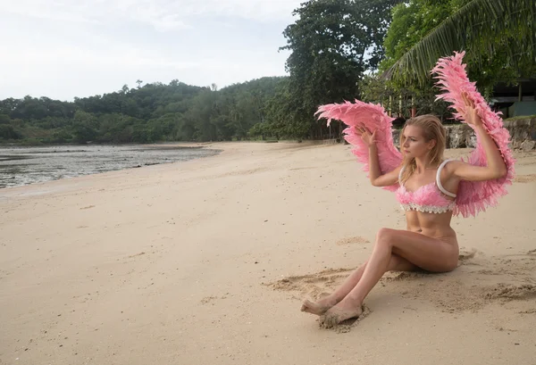 Romantic young beauty as an angel on the tropical beach. Blonde pretty female woman wearing pink wings and feather bra sitting on sandy beach close the sea and looking somewhere over sky and green trees background — Stock Photo, Image