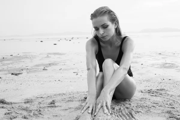 Black and white photo of elegant blonde pretty woman with wet look wearing black swimsuit sitting on the sandy beach near the sea over sky background — Stock Photo, Image