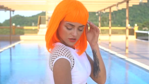 Closeup portret van sexy mooie vrouw in de moderne futuristische stijl poseren bij het dakzwembad op het tijdens zonnige zomerdag. Creatieve blik van getatoeëerd vrouw dragen witte bikini en oranje pruik - slow motion video — Stockvideo