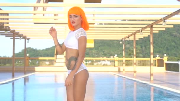 Mujer hermosa atractiva en estilo futurista moderno posando junto a la piscina de la azotea durante el día de verano soleado. Aspecto creativo de mujer tatuada con bikini blanco y peluca naranja - video en cámara lenta — Vídeos de Stock