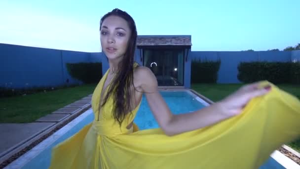 Πανέμορφη γυναίκα της μόδας με σκούρα μαλλιά σε κομψό κίτρινο φόρεμα χαμογελώντας και χορεύοντας δίπλα στην πισίνα σε πολυτελής βίλα-βίντεο σε αργή κίνηση — Αρχείο Βίντεο
